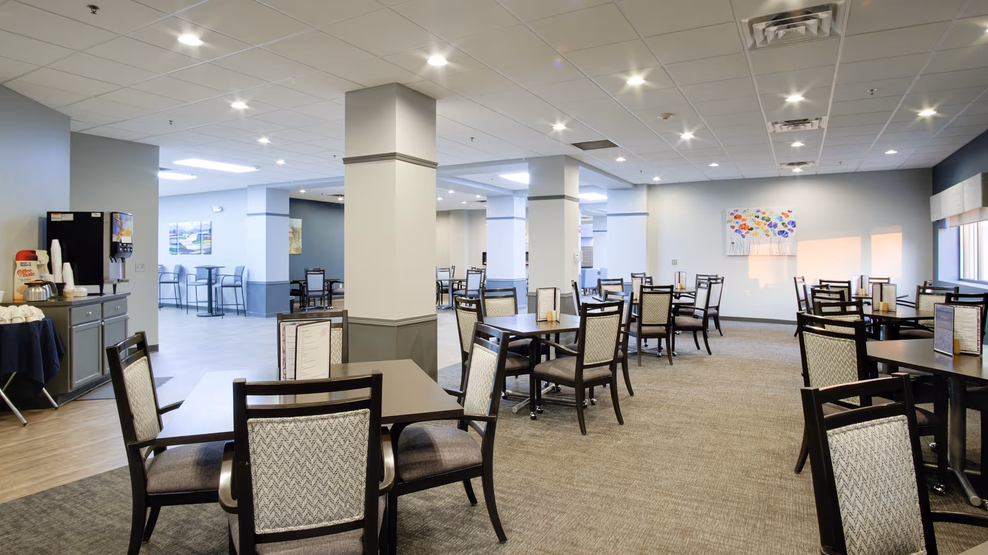 A spacious dining room in a senior living facility with multiple dark wood tables and cushioned chairs arranged neatly. The room has light-colored walls, recessed ceiling lights, and a carpeted floor. There is a beverage station with a coffee maker and cups on the left side, and colorful artwork on the walls. Large windows allow natural light to brighten the space.