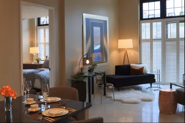 A cozy interior space featuring a small dining area with a table set for two, a living room with a black sofa, a floor lamp, a small side table, and a fluffy white rug. In the background, there is a bedroom visible through an open doorway, with a bed, bedside table, and lamp. Large windows with white shutters allow natural light to fill the room.