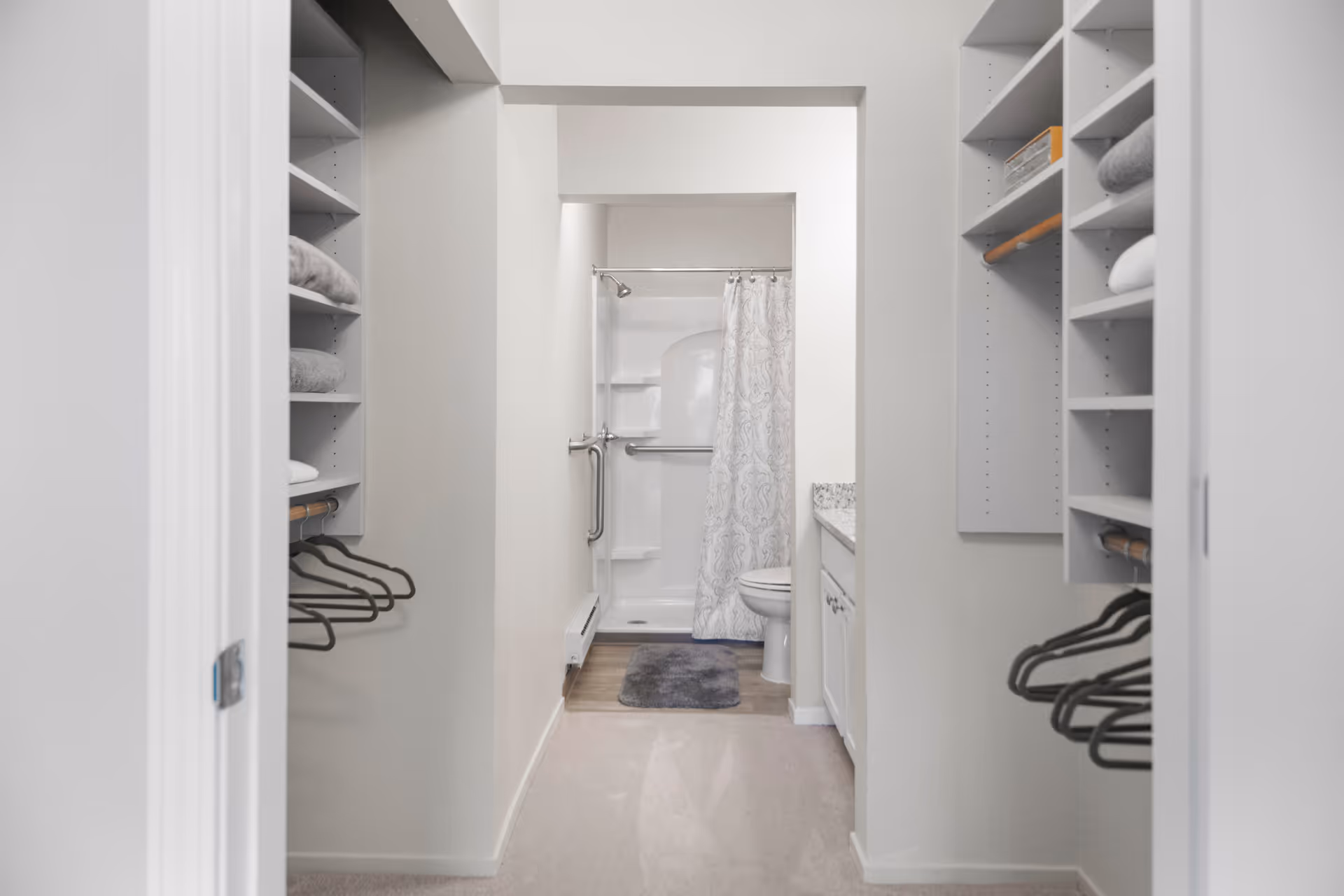 View through a walk-in closet with shelves and hanging rods on both sides, leading to a bathroom with a shower, toilet, and a countertop with cabinets. The bathroom has a white shower curtain with a subtle pattern and a gray bath mat on the floor.