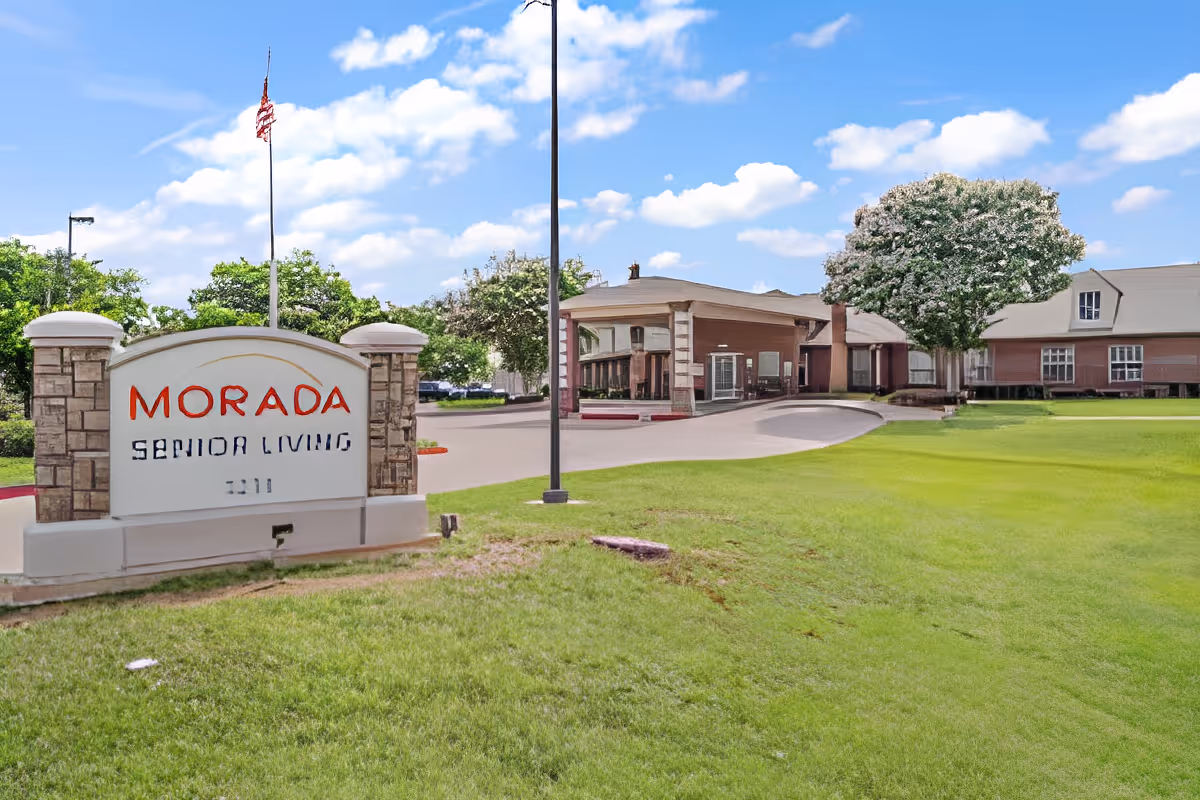 Exterior view of Morada Senior Living facility with a large sign in the foreground displaying the facility name. The building is surrounded by green grass, trees, and a clear blue sky with some clouds.