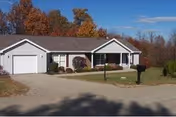 Single-story residential building with a garage, front porch, and mailbox, surrounded by grass and trees with autumn foliage under a clear blue sky.