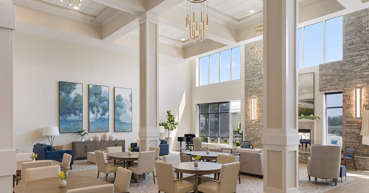 Spacious sunlit communal lounge with high ceilings, round dining tables, seating areas, and a stone fireplace.