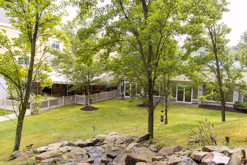 Grassy courtyard with trees, a rock-lined water feature, and the exterior of a senior residence with a covered porch and railing.