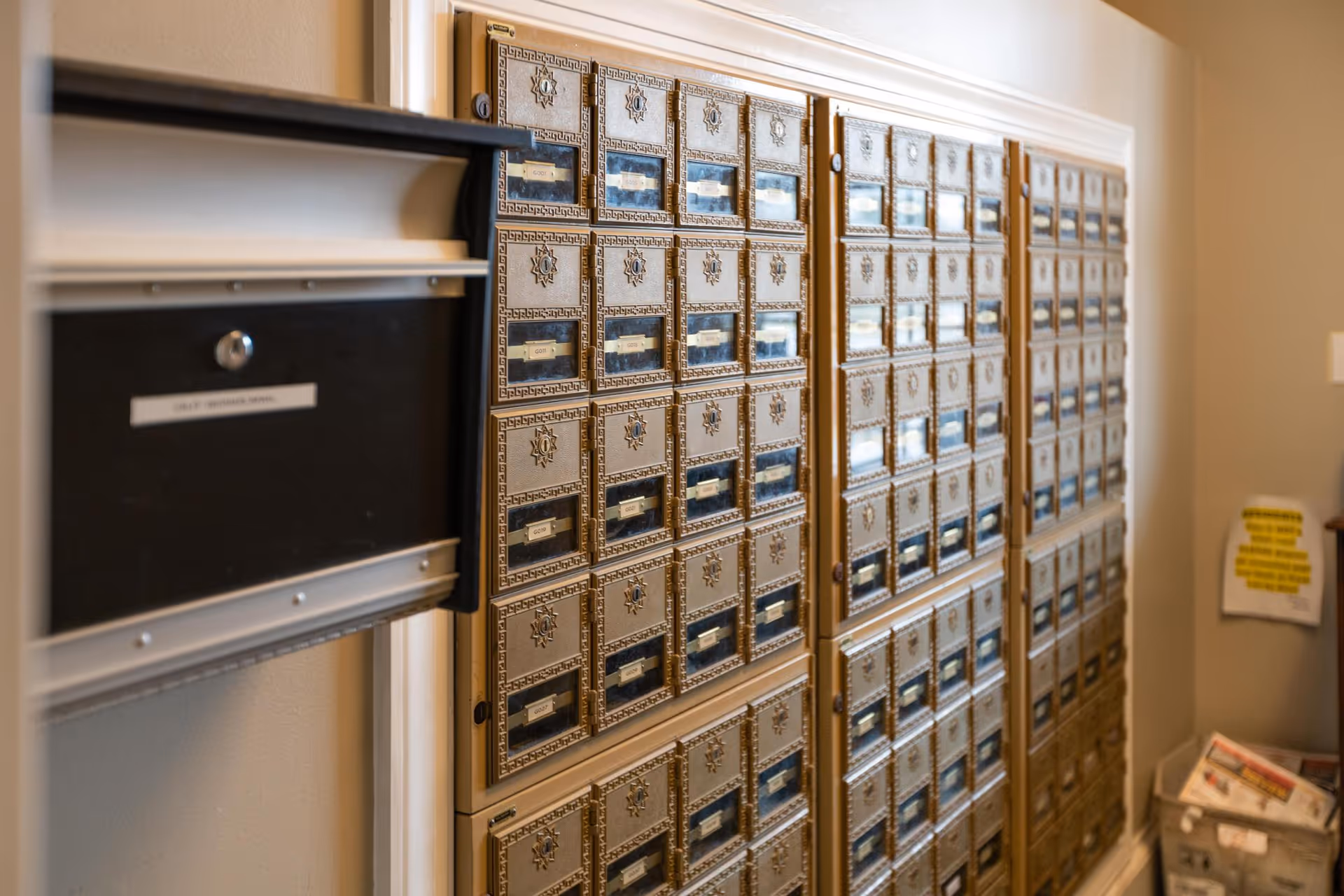A wall of vintage-style mailboxes with multiple small locked compartments, each with a label holder and a decorative star emblem. To the left, there is a larger black mailbox labeled 'Staff Outgoing Mail.' In the background, there is a bin with newspapers and a wall with a posted notice.