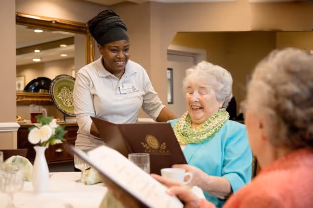 A smiling staff member in a white polo shirt assists two elderly women seated at a dining table in a warm, well-lit room. One elderly woman, wearing a turquoise top and a green scarf, is looking at a menu and smiling, while the other woman, in an orange top, is also holding a menu. The table is set with glasses, a cup, and a small vase with white flowers.