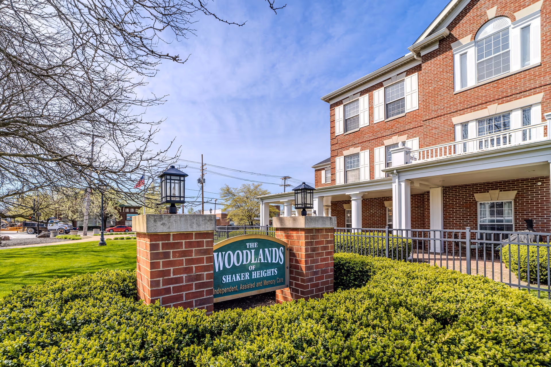 Exterior view of The Woodlands of Shaker Heights senior living facility showing a brick building with white trim and multiple windows. In the foreground, there is a green sign with white text mounted between two brick pillars with lanterns on top. The area is surrounded by green bushes and a lawn, with a clear blue sky above.