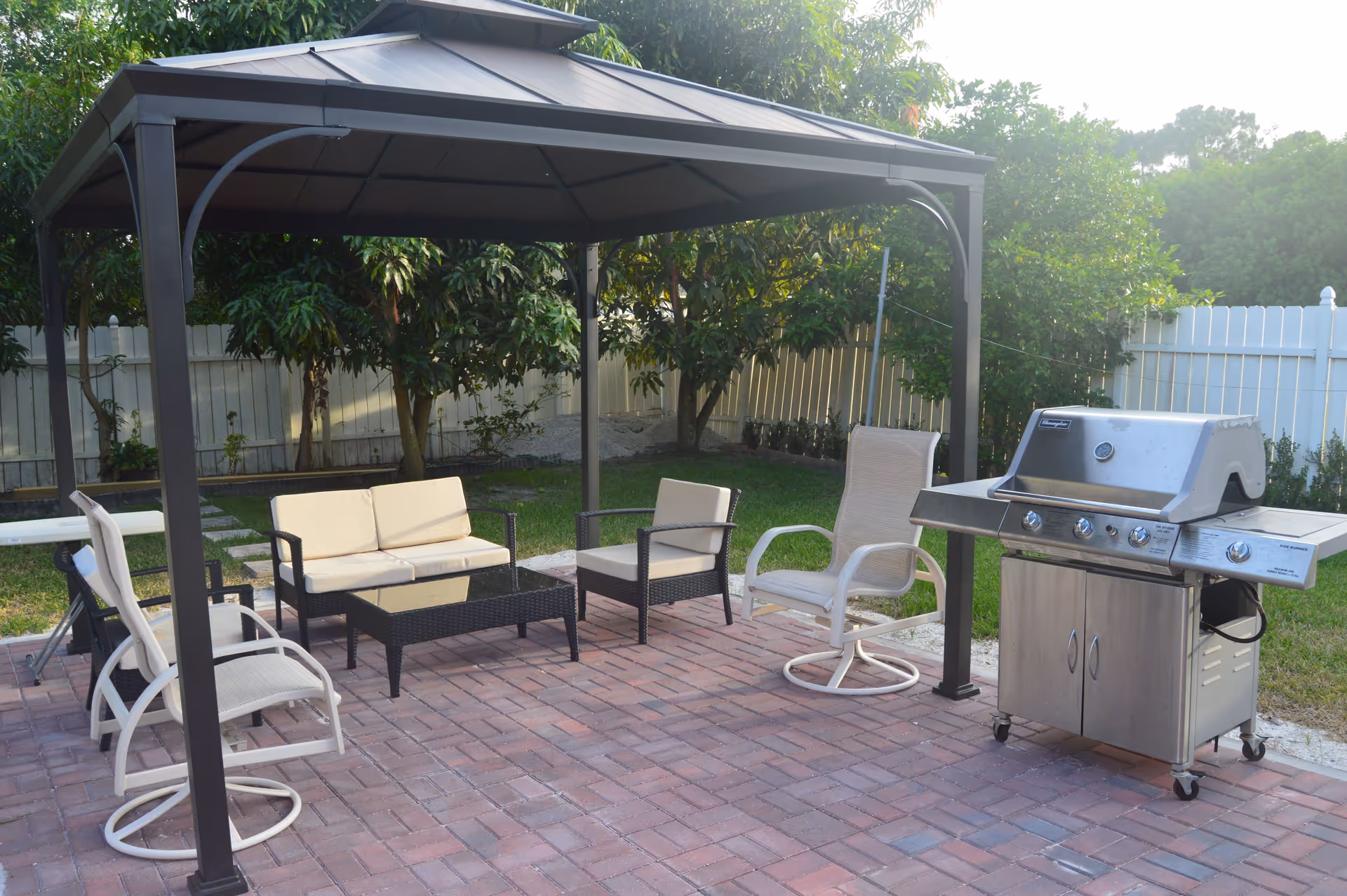 Outdoor patio area with a metal gazebo covering cushioned seating including a loveseat, two armchairs, and a glass-top coffee table. There is a stainless steel gas grill on the right side, and the area is surrounded by green grass, trees, and a white fence.