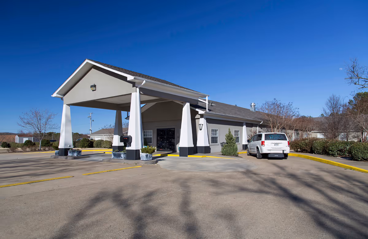 Exterior view of Quapaw Nursing & Rehab Center showing the main entrance with a covered drop-off area supported by white pillars. A white van is parked near the entrance, and the surrounding area includes bushes, trees, and a clear blue sky.