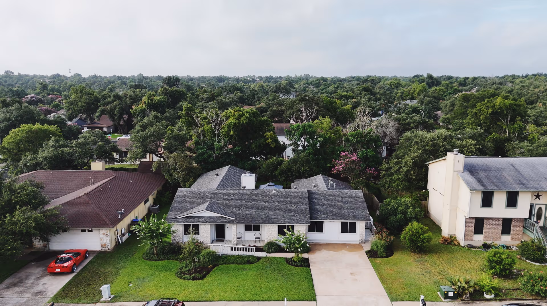 Aerial view of a residential neighborhood showing three houses with well-maintained lawns and driveways, surrounded by dense green trees under a cloudy sky.