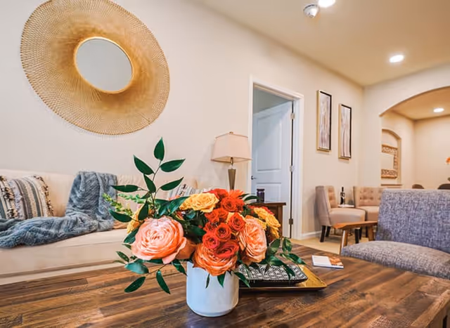 A cozy living room area with a wooden coffee table in the foreground holding a white vase filled with colorful flowers including orange, pink, and red roses. Behind the table, there is a beige sofa with patterned cushions and a blue throw blanket. A round decorative mirror with a golden frame hangs on the wall above the sofa. To the right, there is a gray upholstered chair and a small wooden side table with a lamp. The room has light-colored walls and wooden flooring, with an open doorway and framed artwork visible in the background.