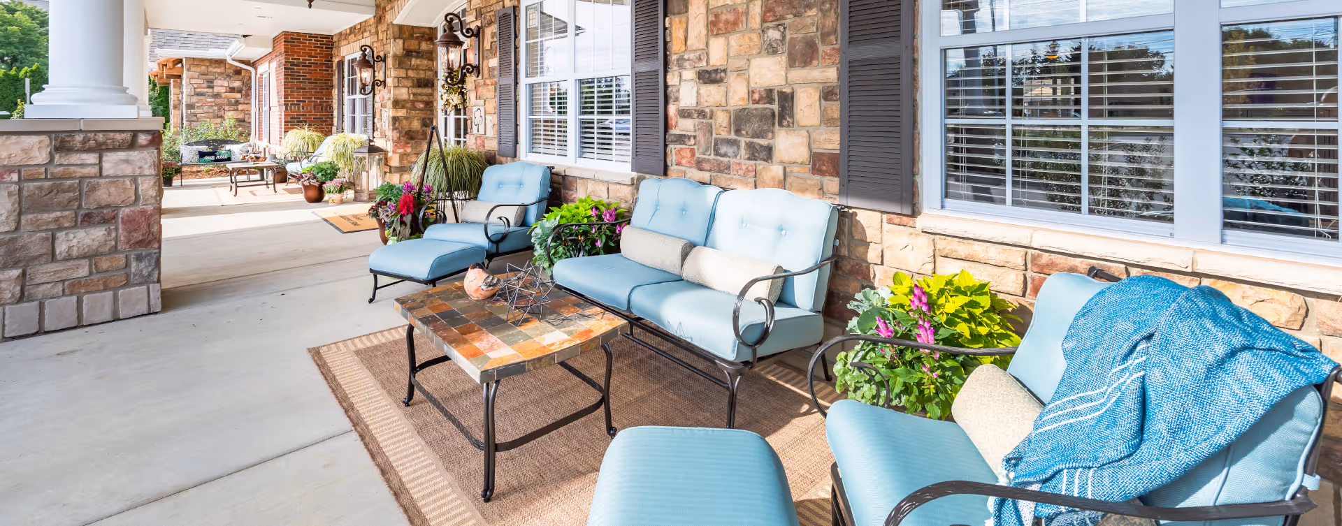 Outdoor patio area with cushioned seating including chairs and a loveseat, a small table with a decorative item, potted plants, and a stone wall with windows and shutters in the background.