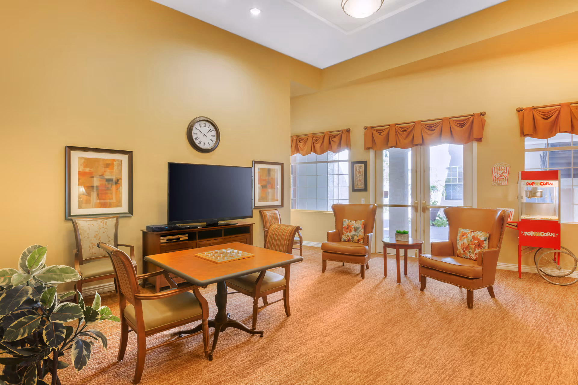 A cozy living room area with beige walls and carpet. The room features a wooden table with four chairs, a flat-screen TV mounted on a wooden stand, two armchairs with floral cushions, a small round side table, and a red popcorn machine near the window. There are three windows with orange valances letting in natural light, and a clock hangs on the wall above the TV.