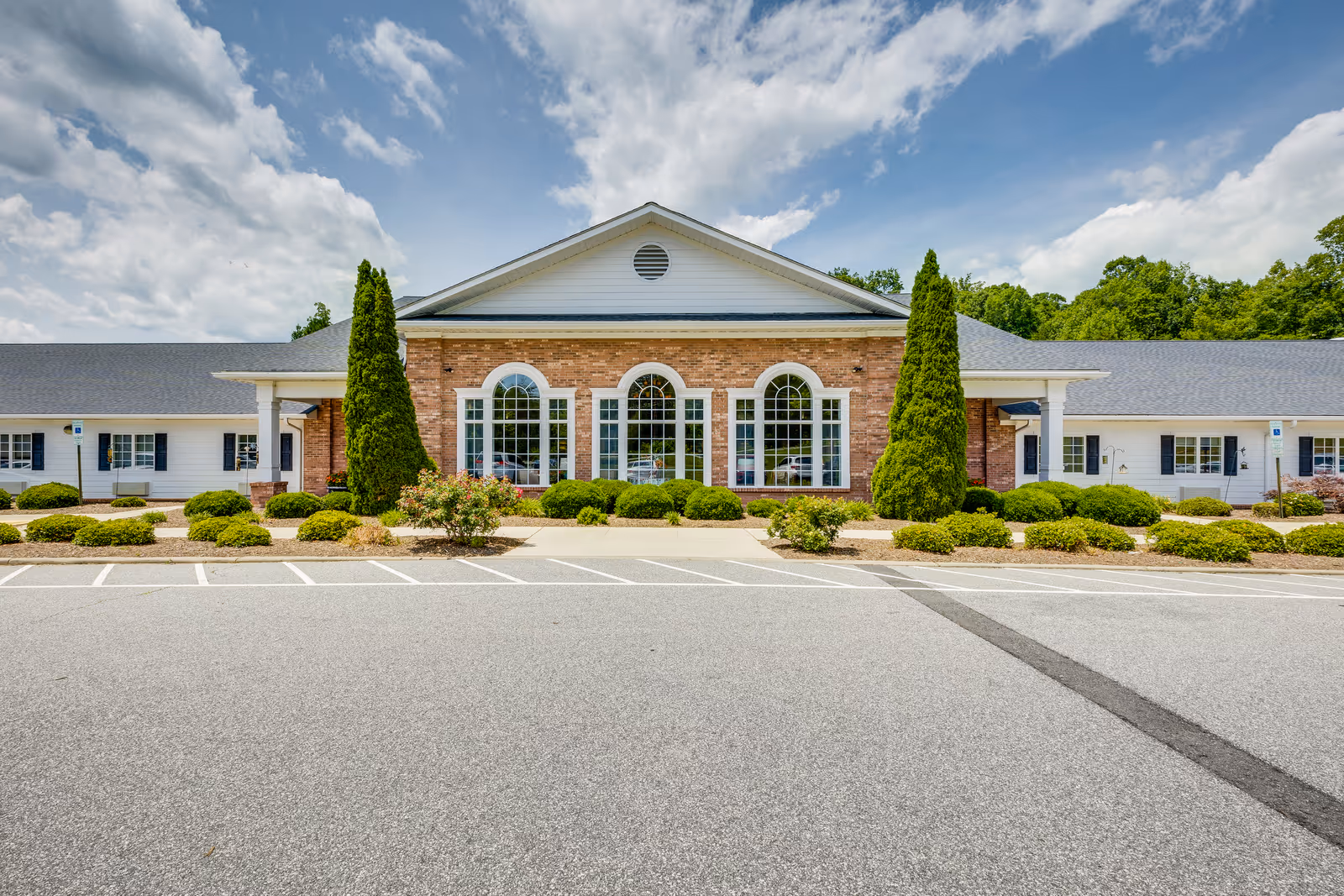 Front exterior view of Priddy Manor Assisted Living & Memory Care building with a brick facade and large arched windows, surrounded by neatly trimmed bushes and two tall evergreen trees, under a partly cloudy sky.