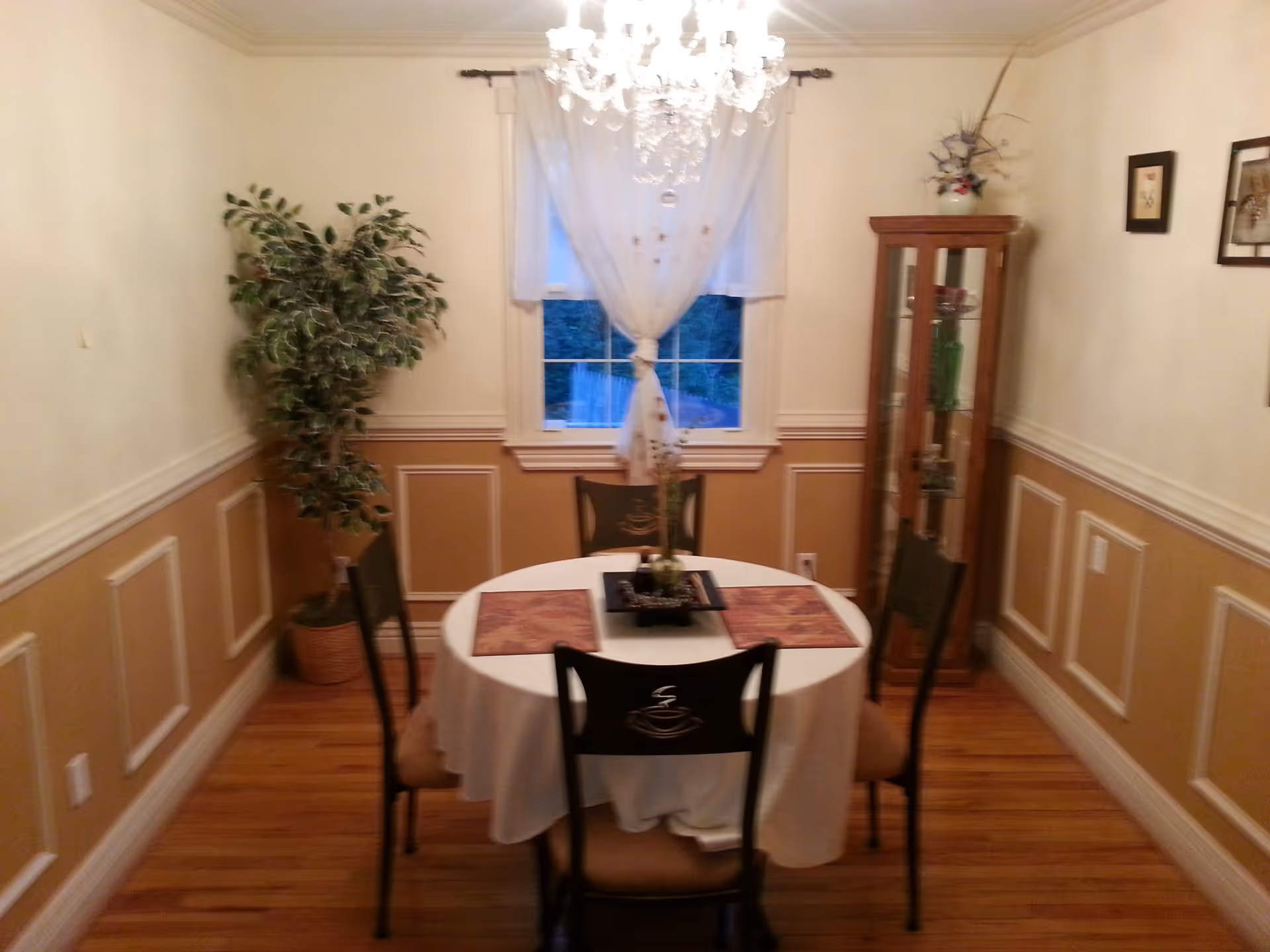 A cozy dining room with a round table covered with a white tablecloth and four chairs around it. The table has placemats and a centerpiece. There is a large window with sheer white curtains, a chandelier hanging from the ceiling, a tall potted plant in the corner, and a glass display cabinet with decorative items. The walls are painted beige with white trim and wainscoting, and the floor is wooden.
