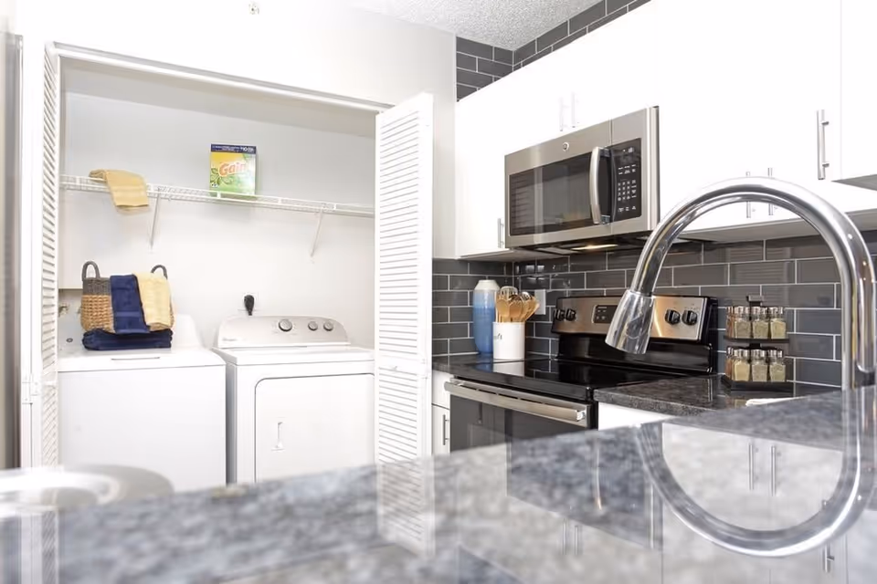 Modern kitchen with white cabinets, black granite countertops, a stainless steel microwave, stove, and dishwasher. To the left, there is a laundry area with a washer and dryer behind white louvered doors, with a shelf above holding a box of Gain detergent and some folded towels.