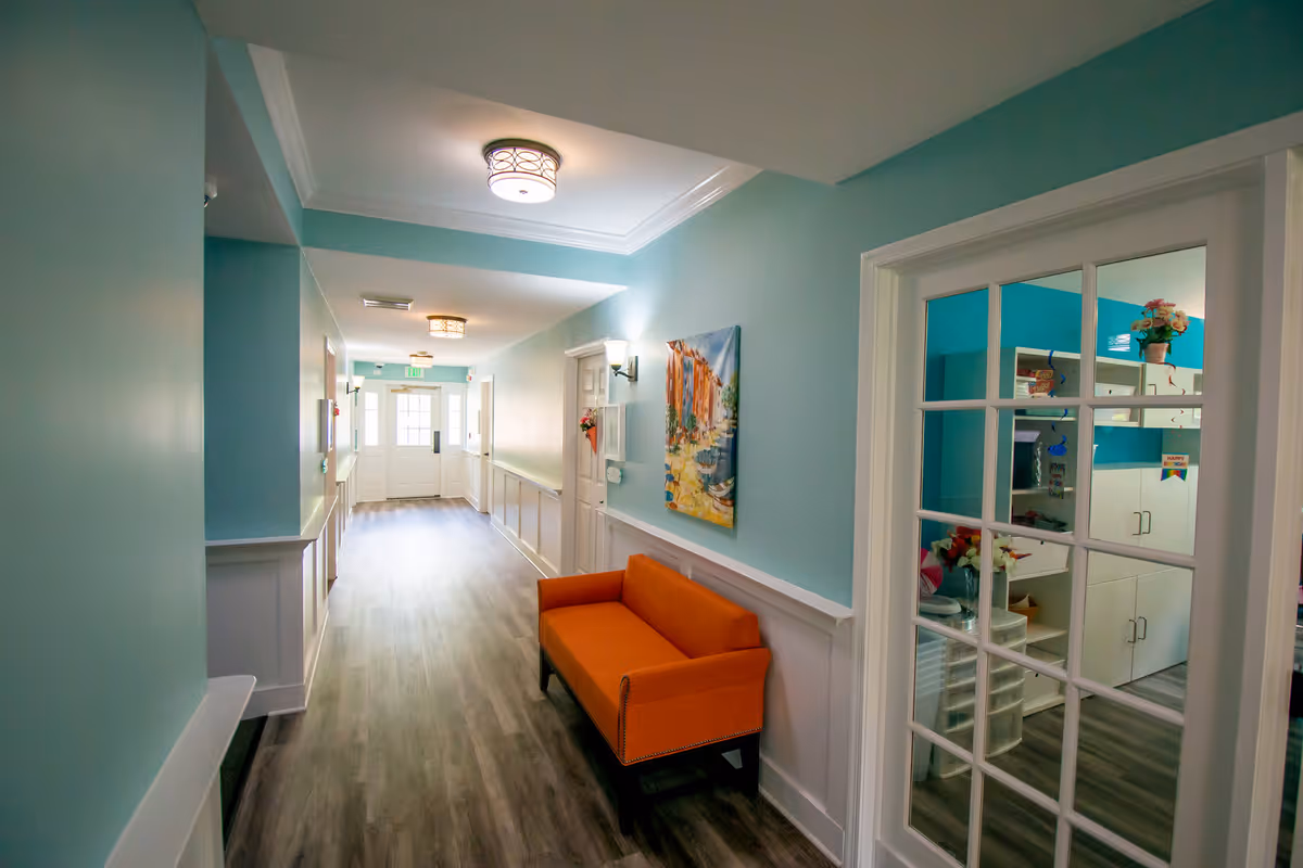 A brightly lit hallway with light blue walls and wood flooring. There is an orange bench along the right wall beneath a colorful painting. The hallway leads to a door with windows letting in natural light. On the right side, there is a glass-paneled door revealing a room with white cabinets, a vase of flowers, and birthday decorations.