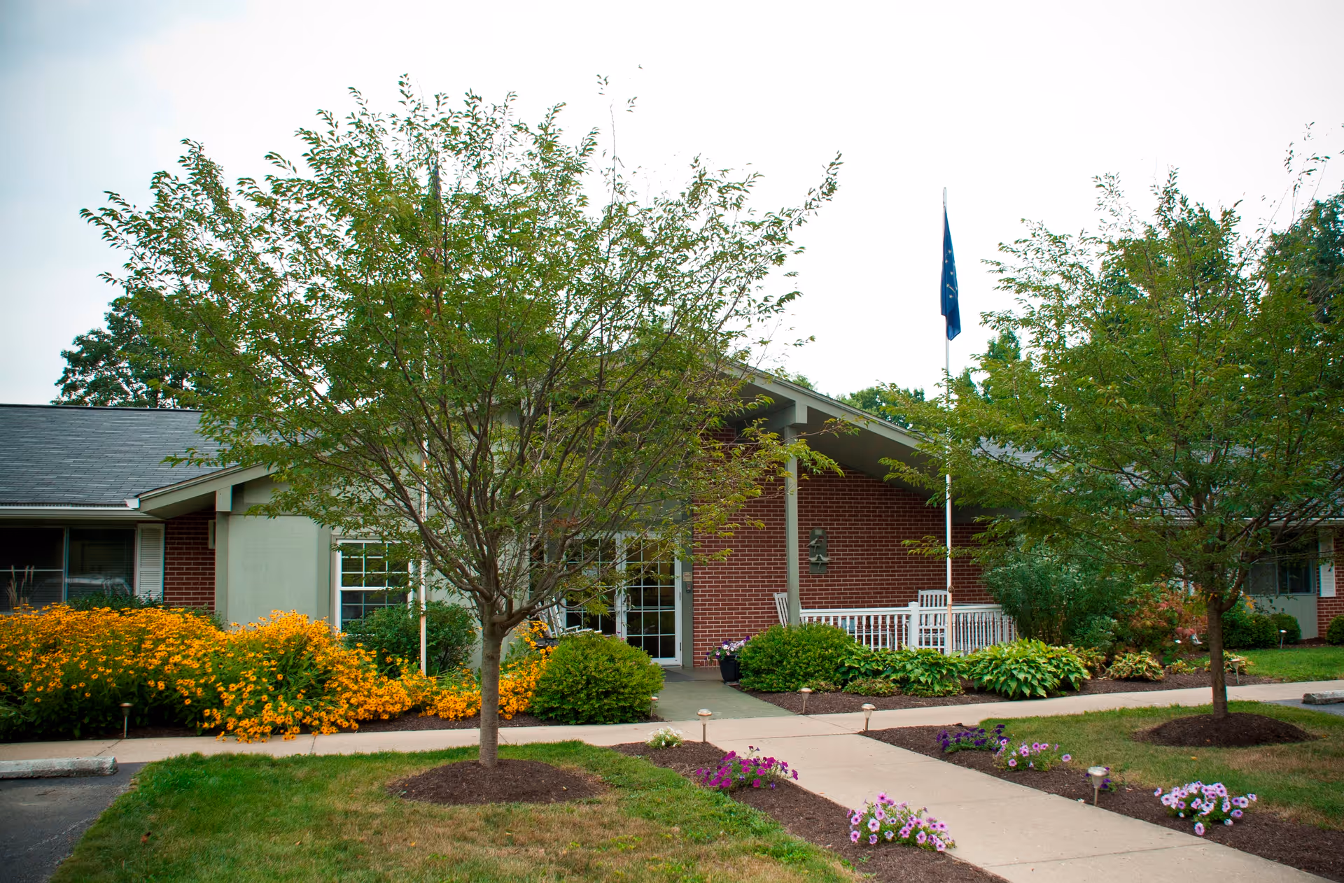 Front entrance of a brick senior living building with a walkway, trees, flowerbeds, and a flagpole.