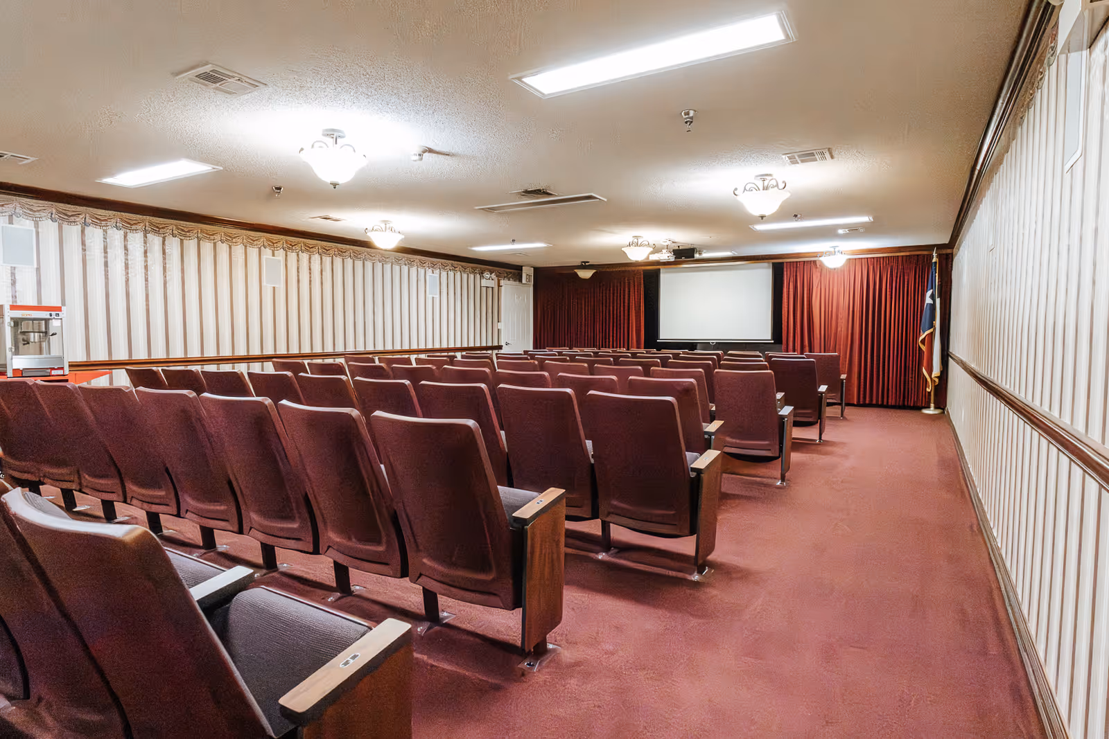 An empty auditorium or theater room with rows of maroon cushioned seats facing a large projection screen at the front. The room has beige walls with vertical striped wallpaper, red carpet flooring, ceiling lights, and a popcorn machine on the left side near the wall.