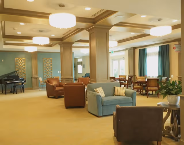 A spacious, well-lit common area with beige carpeting and a coffered ceiling with recessed lighting and hanging light fixtures. The room features a variety of seating including a blue loveseat with striped pillows, brown leather armchairs, and a dark brown chair near a small round table with a potted plant. In the background, there are wooden dining tables and chairs near large windows with teal curtains. A black grand piano is visible on the left side of the room.