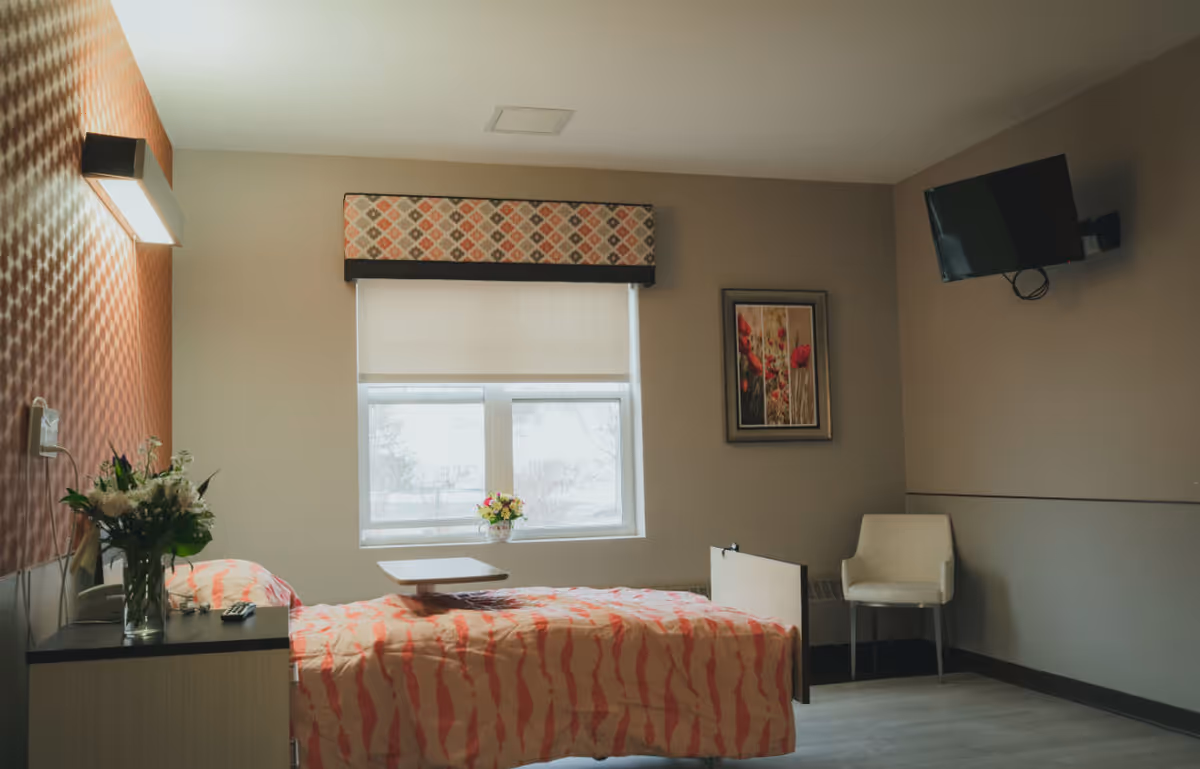 A single bed with a coral and beige patterned bedspread in a room with beige walls. There is a small table over the bed, a vase with flowers on a bedside cabinet, a window with a patterned valance and a white blind, a framed picture of red flowers on the wall, a white chair in the corner, and a wall-mounted flat screen TV.