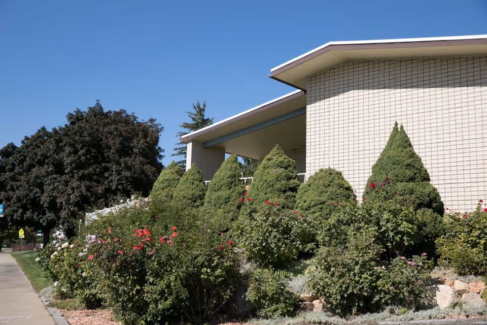 Exterior view of a building with a beige tiled wall and a covered entrance. In front of the building, there are neatly trimmed conical evergreen bushes and flowering plants with red and pink blooms. A sidewalk runs alongside the garden area under a clear blue sky.
