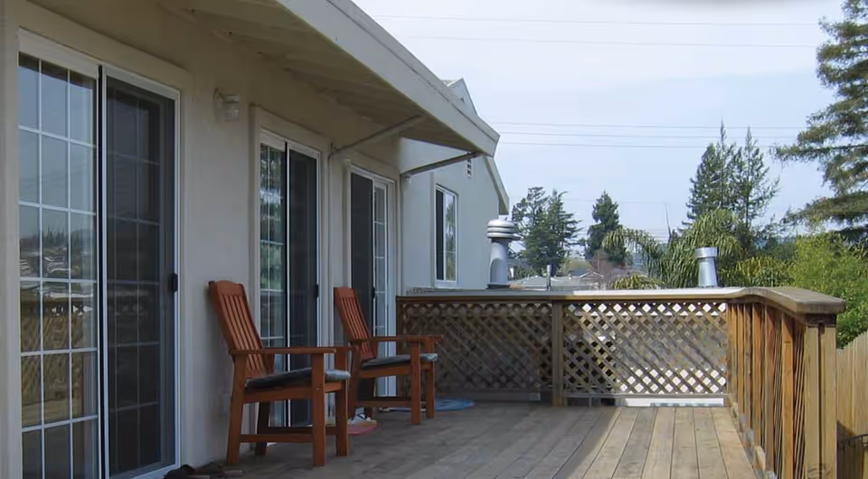 Wooden deck attached to a building with two wooden chairs placed near sliding glass doors. The deck has a wooden railing with lattice panels and overlooks trees and rooftops in the distance under a clear sky.