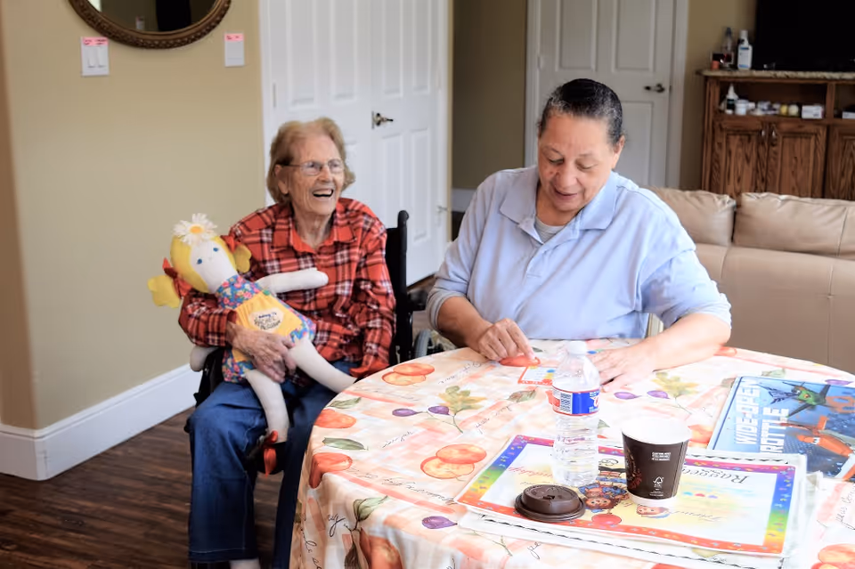An elderly woman in a red plaid shirt sitting in a wheelchair holding a doll, smiling and looking at a caregiver in a light blue shirt who is sitting at a table covered with a fruit-patterned tablecloth. The caregiver is engaged with some papers on the table, which also has a water bottle, a coffee cup, and a children's book. The room has wooden flooring, beige walls, a round mirror, and a beige couch in the background.