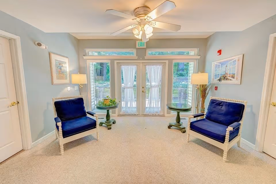 A bright and cozy sitting area with two blue cushioned armchairs facing each other, each accompanied by a small round green table. The room has light blue walls, beige carpet, two floor lamps, framed artwork on the walls, and a ceiling fan with lights. Large glass double doors with sheer white curtains lead to an outdoor area with greenery visible outside.