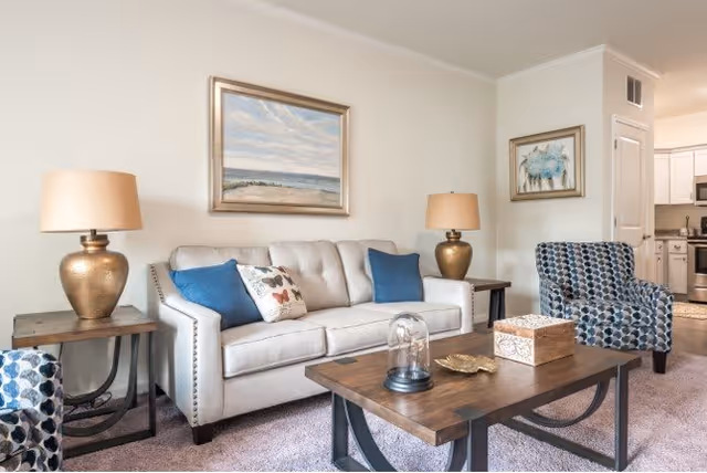 A bright and cozy living room featuring a beige sofa with blue and patterned throw pillows, two patterned armchairs, two wooden side tables with matching gold lamps, a wooden coffee table with decorative items, and framed artwork on the walls. The room has light-colored walls and carpeted flooring, with a partial view into a kitchen area.