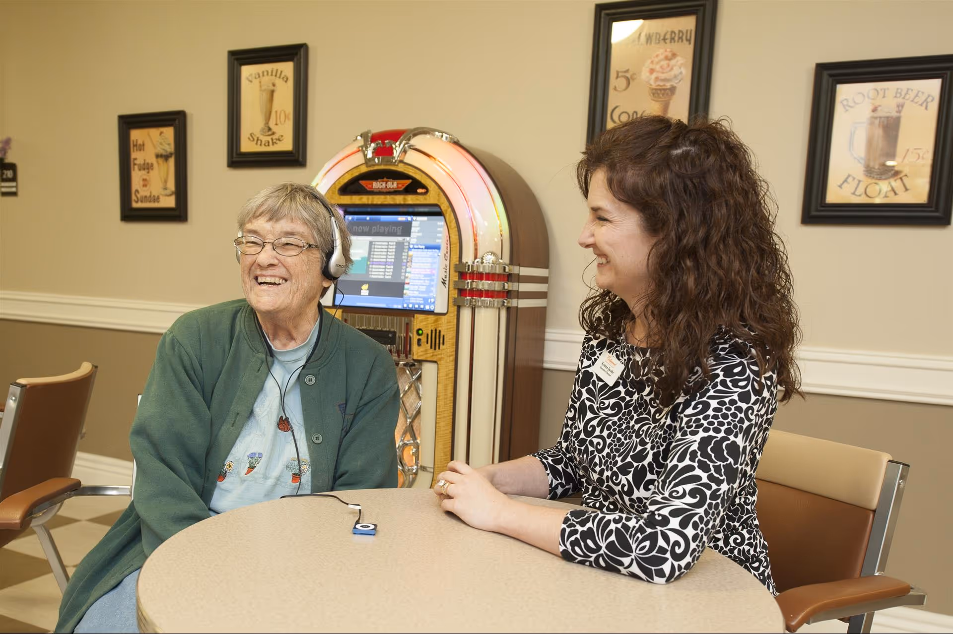 An elderly woman wearing headphones and a green jacket sits at a round table smiling, while a woman with curly hair and a black and white patterned top sits across from her, also smiling. Behind them is a vintage-style jukebox and framed retro posters on the wall advertising vanilla shakes, hot fudge sundaes, strawberry cones, and root beer floats.