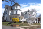 Two white multi-story Victorian-style houses with porches and front steps on a small lawn under a blue sky.