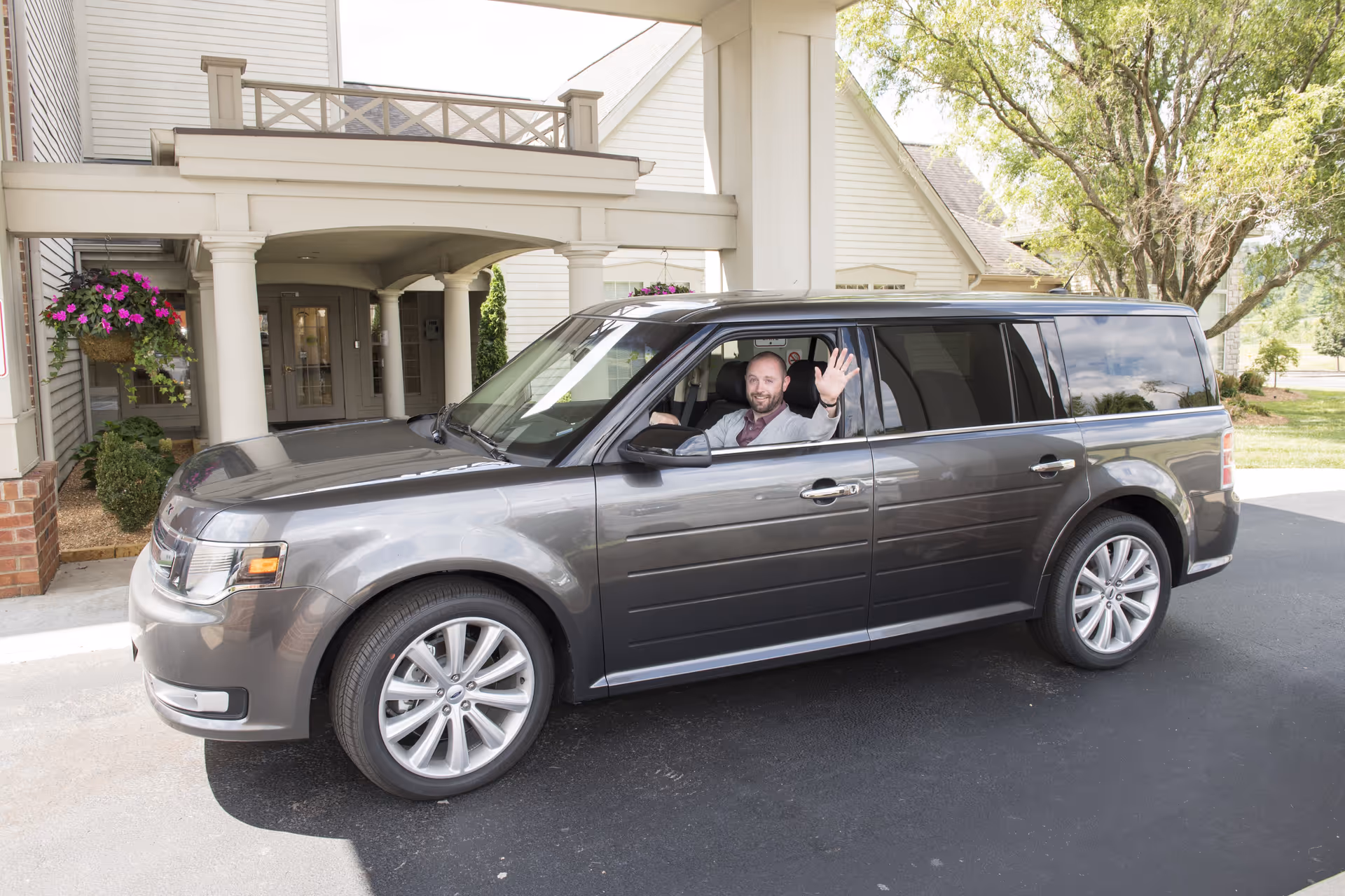A man sitting in the driver's seat of a gray SUV parked outside the entrance of a building with white columns and hanging flower baskets. The man is smiling and waving.