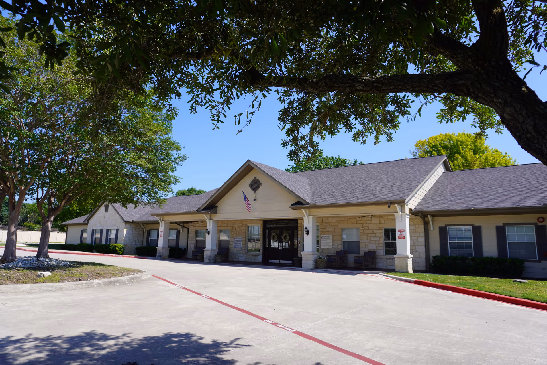 Exterior view of a single-story senior living facility building with a stone facade and a covered entrance. There are trees providing shade in the foreground and an American flag hanging near the entrance. The sky is clear and blue.