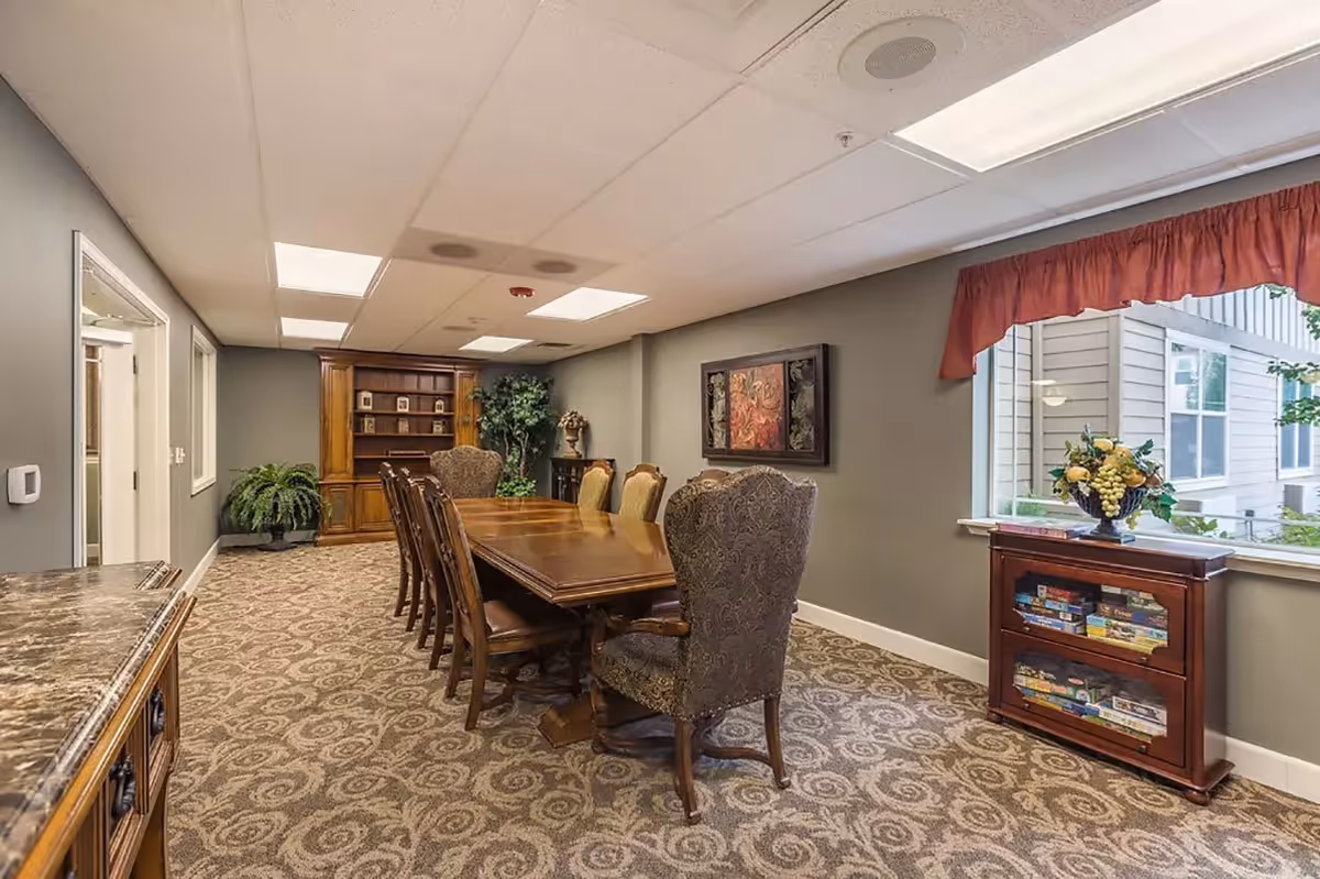 A formal dining room with a long wooden table surrounded by eight upholstered chairs. The room has patterned carpet, gray walls, and a large window with a red valance. There is a wooden cabinet with board games and a decorative fruit arrangement under the window, and a wooden bookshelf and plants at the far end of the room.