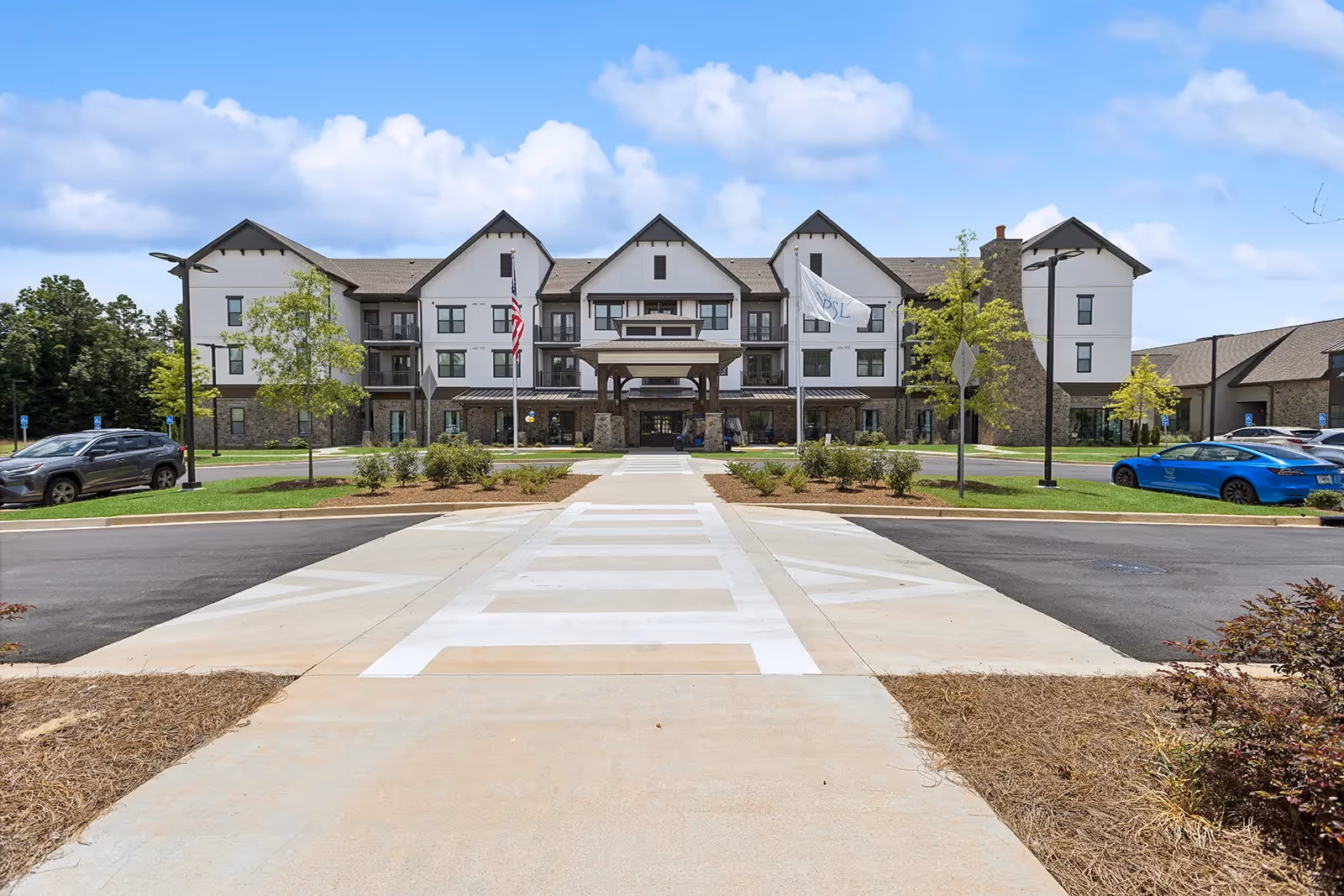 Front exterior view of The Phoenix at Opelika, a multi-story senior living facility with a covered entrance, landscaped grounds, and parked cars on either side of the driveway under a partly cloudy sky.