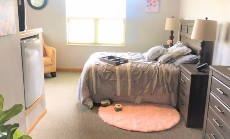 A cozy bedroom with a bed covered in a beige comforter and multiple pillows. There is a small pink circular rug on the floor near the foot of the bed, with two small pet beds on it. A wooden nightstand with a lamp and a phone sits next to the bed, and a matching dresser is visible on the right. A yellow armchair is placed near a window with blinds, and a small refrigerator is built into the wall on the left side. The room has neutral-colored walls and carpeted flooring.