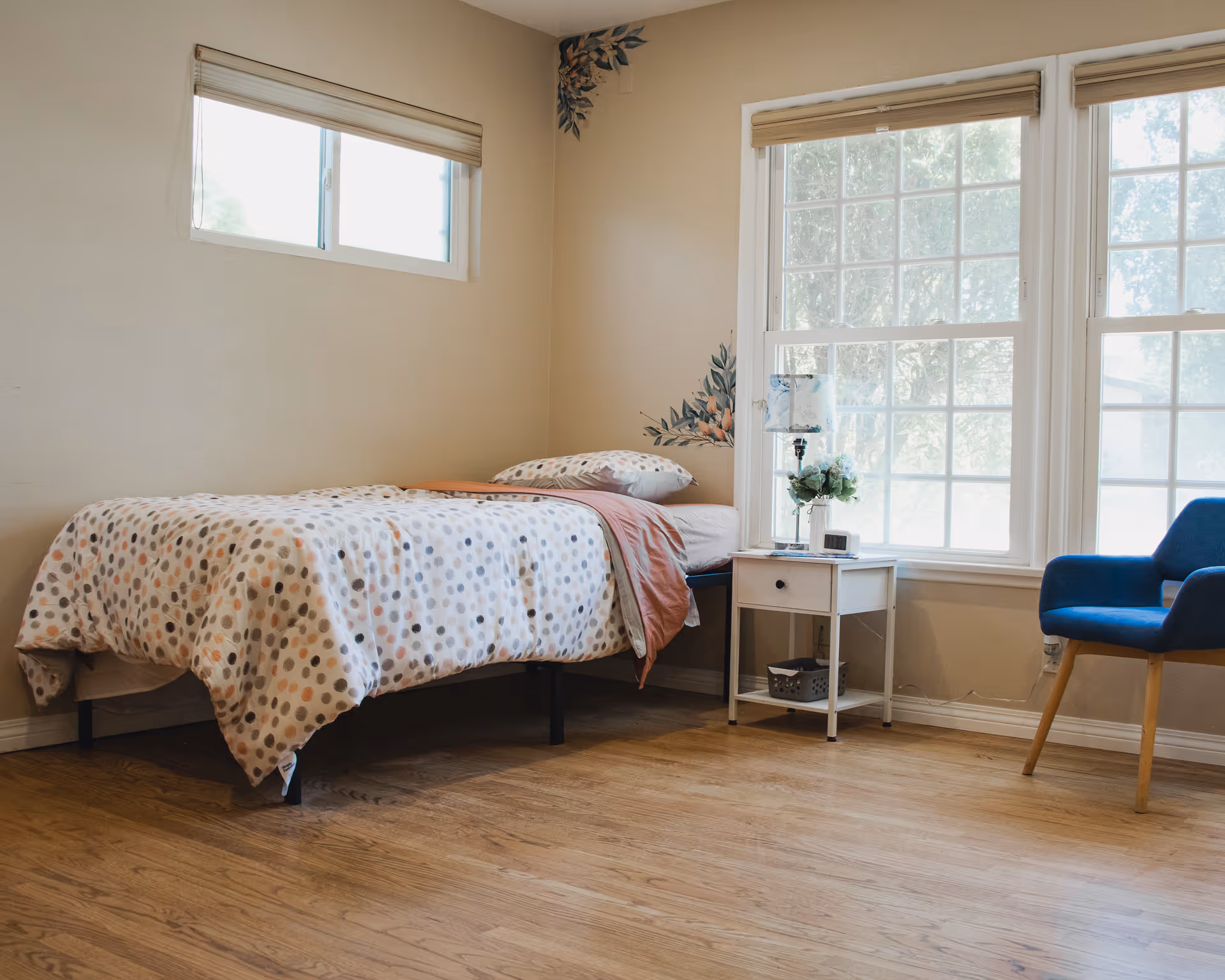 A bright and cozy bedroom with a single bed covered in a polka dot patterned duvet and a pink throw blanket. Next to the bed is a white nightstand with a lamp, a small plant, and a clock. There are two large windows with beige blinds letting in natural light, and a blue upholstered chair with wooden legs is placed near the window. The walls are beige with a floral corner decal, and the floor is wooden.