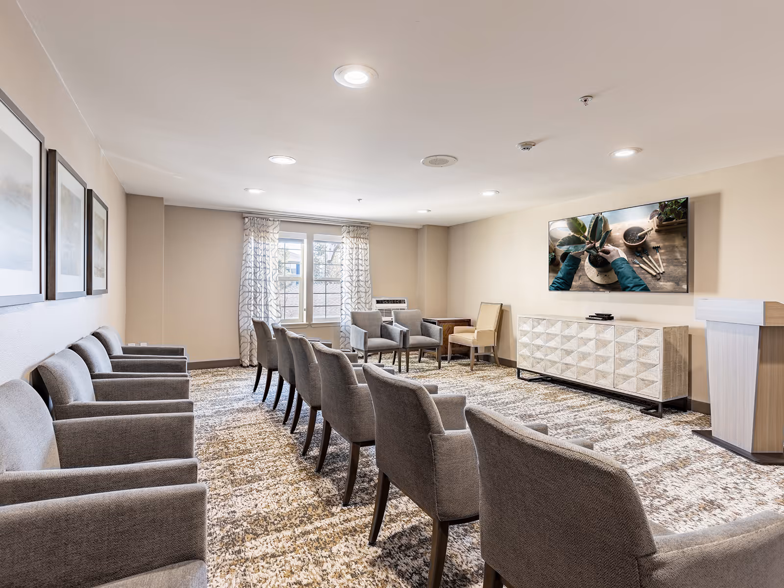 A well-lit meeting or presentation room with rows of gray upholstered chairs arranged facing a large flat-screen TV mounted on the wall. The room has beige walls, patterned carpet, framed artwork on one wall, a window with patterned curtains, and a light-colored wooden podium and cabinet beneath the TV.