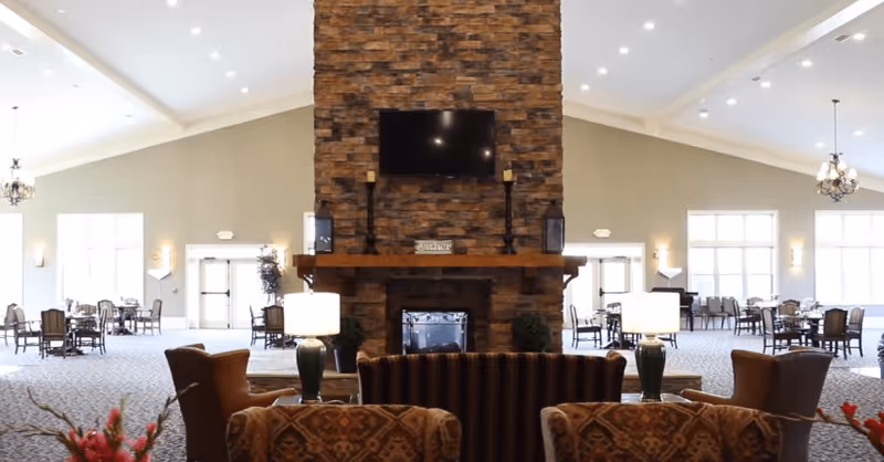 Spacious senior living common area with a large stone fireplace in the center, a mounted TV above it, and comfortable seating including armchairs and sofas arranged around the fireplace. The room has high vaulted ceilings with chandeliers and multiple tables and chairs near large windows allowing natural light.