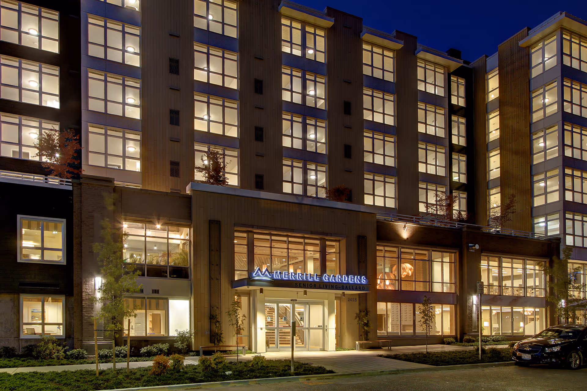 Exterior nighttime view of Merrill Gardens at Ballard senior living facility with illuminated windows and entrance sign, surrounded by landscaping and a parked car.