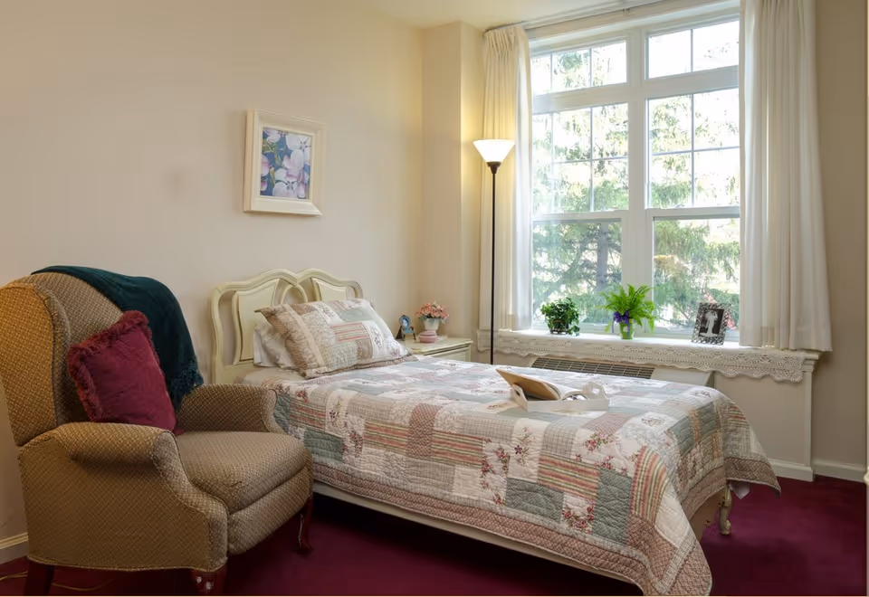A neat bedroom with a single bed covered by a patchwork quilt, an upholstered armchair, a floor lamp, and a large window with potted plants.