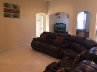 Interior view of a living room with beige walls and tiled floor. The room features two dark brown leather sofas with patterned cushions. There is a flat-screen TV mounted on the wall in an adjacent room visible through an arched doorway. Natural light enters through a window with sheer curtains.
