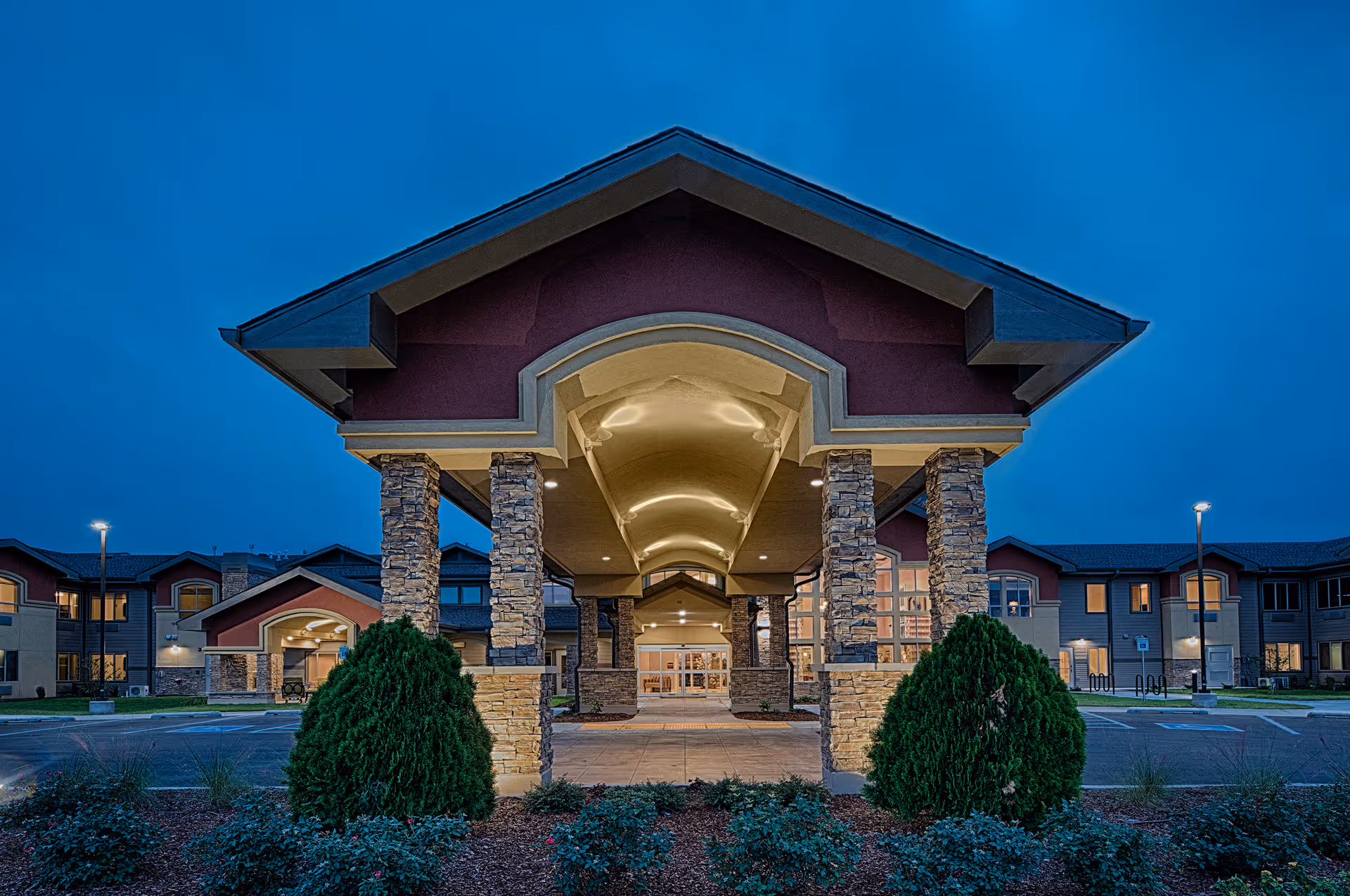 Front entrance of an assisted living building with an illuminated porte-cochere supported by stone columns at dusk.