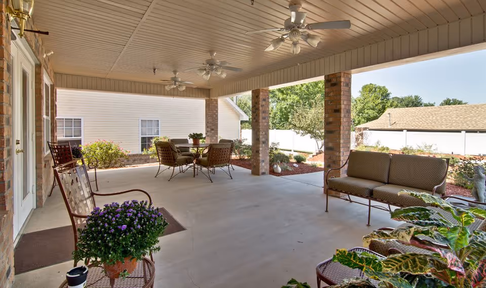 Covered outdoor patio area with ceiling fans, cushioned seating including a sofa and chairs, a round table with chairs, potted plants, and a view of a garden with bushes and trees under a clear sky.