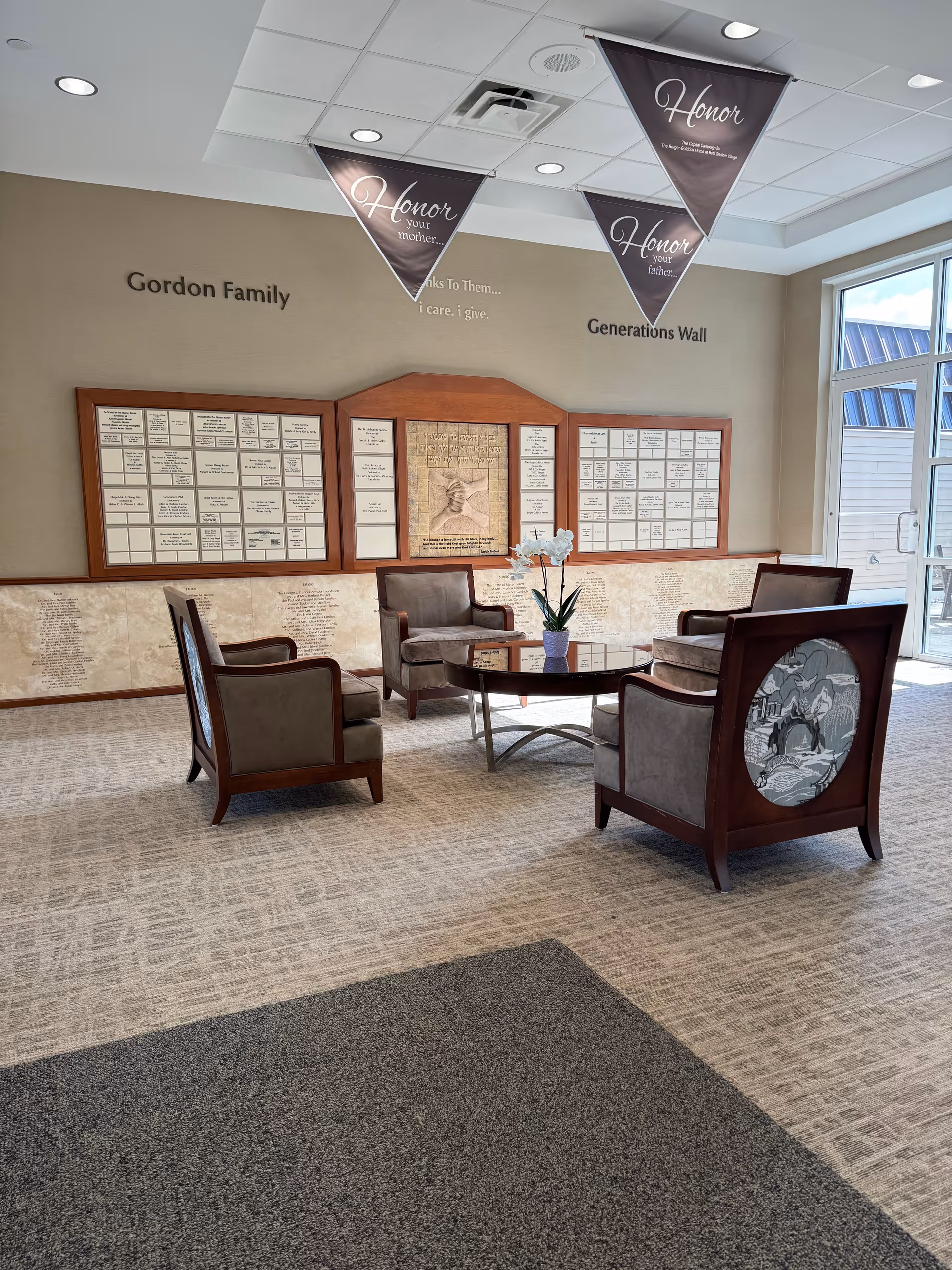 A seating area inside a facility with four cushioned chairs arranged around a circular glass coffee table with a potted orchid on it. Behind the seating area is a wall with plaques and inscriptions titled 'Gordon Family Generations Wall'. Triangular banners hang from the ceiling with the word 'Honor' and phrases like 'Honor your mother' and 'Honor your father'. Large windows on the right side let in natural light.
