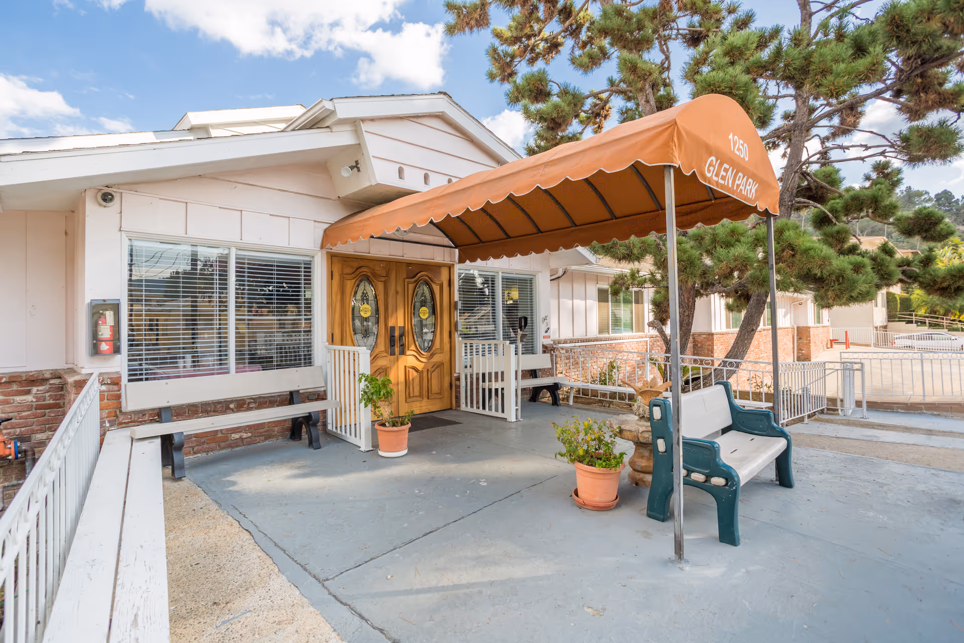Entrance of Glen Park at Glendale - Boynton facility with double wooden doors, a brown awning labeled '1250 GLEN PARK', benches, potted plants, and trees nearby under a partly cloudy sky.