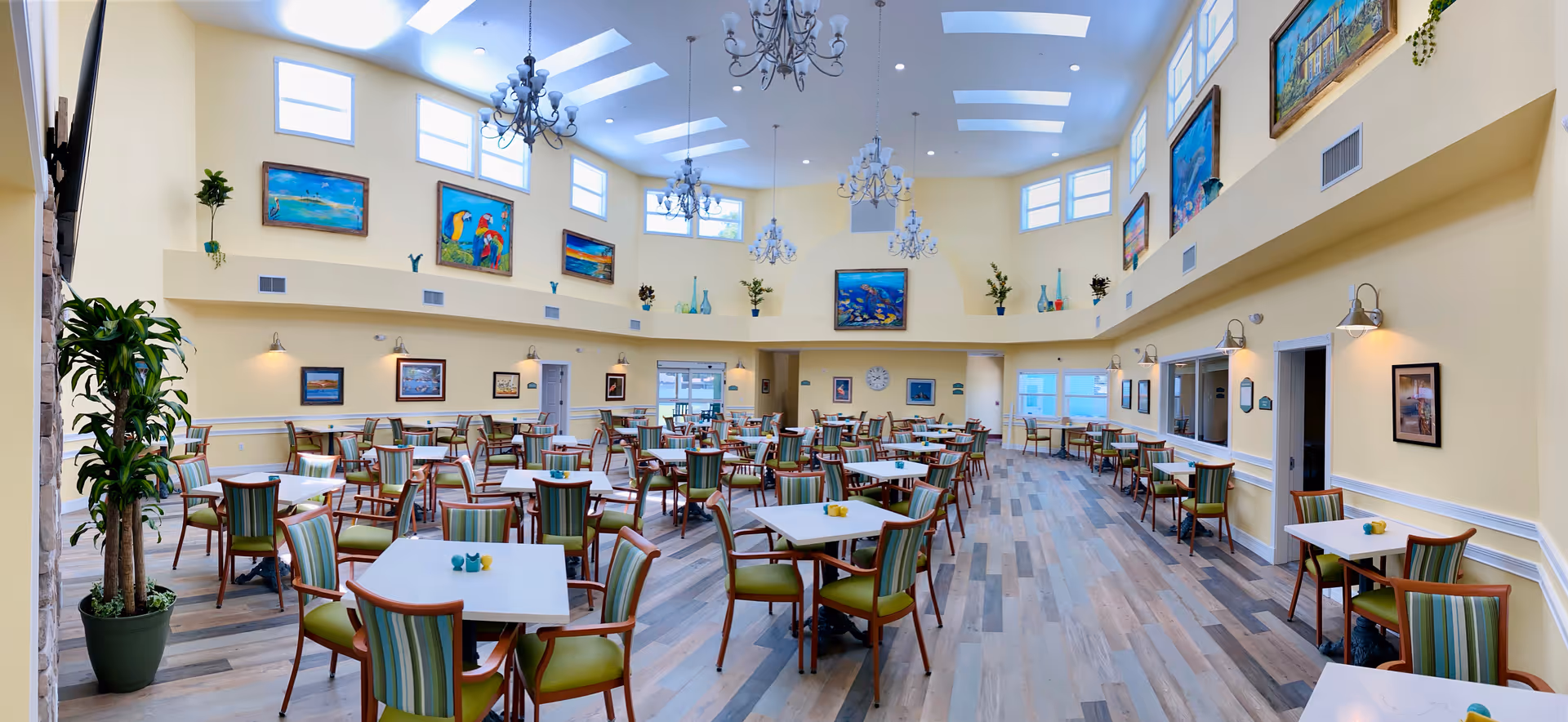 Bright, spacious assisted-living dining room with many tables and chairs, high ceilings, chandeliers, and artwork on the walls.
