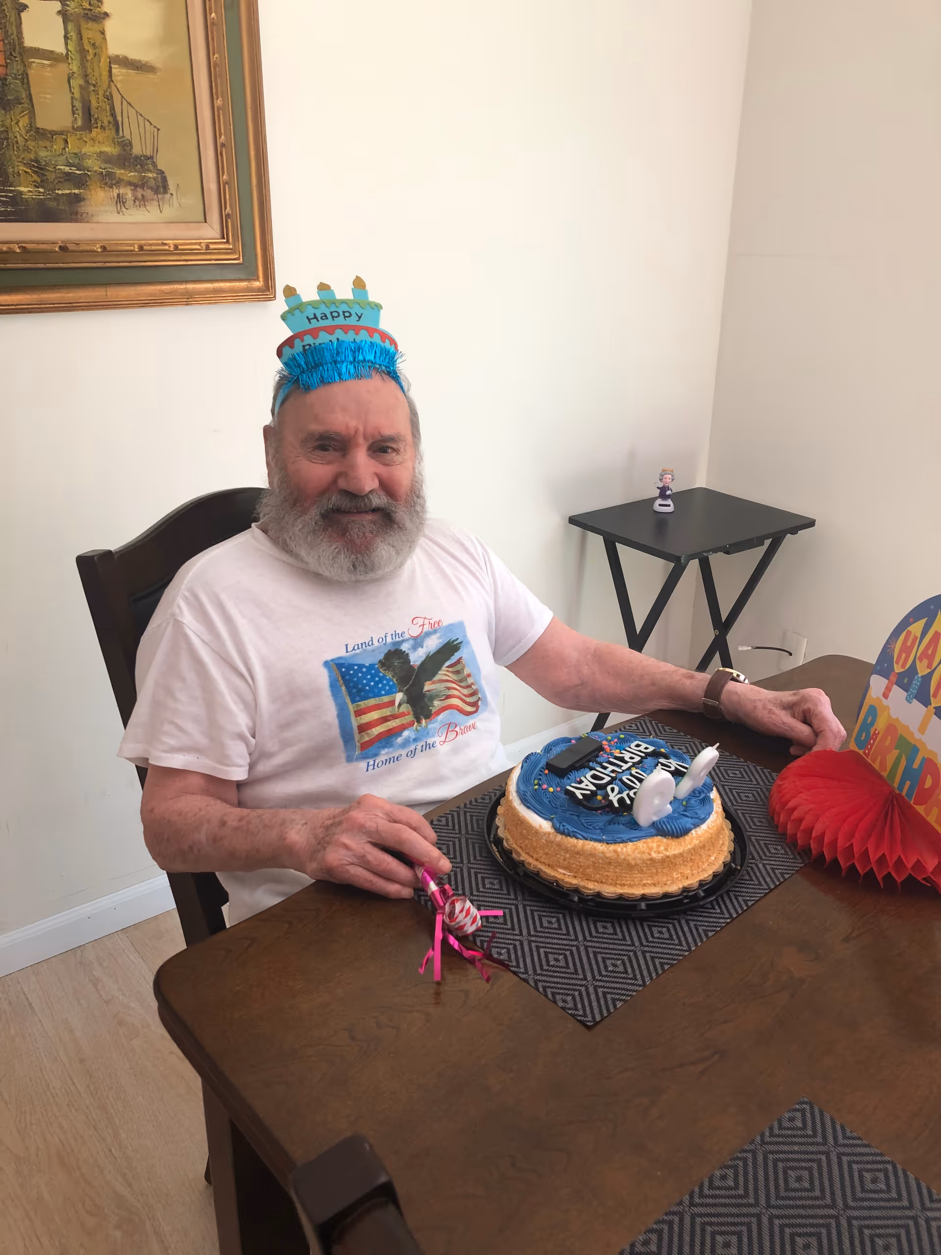 An elderly man with a white beard wearing a white t-shirt and a blue birthday hat sits at a wooden dining table with a blue birthday cake in front of him. The table has a black patterned placemat, and there is a colorful birthday decoration on the table. A small black side table with a figurine is in the background, along with a framed painting on the wall.
