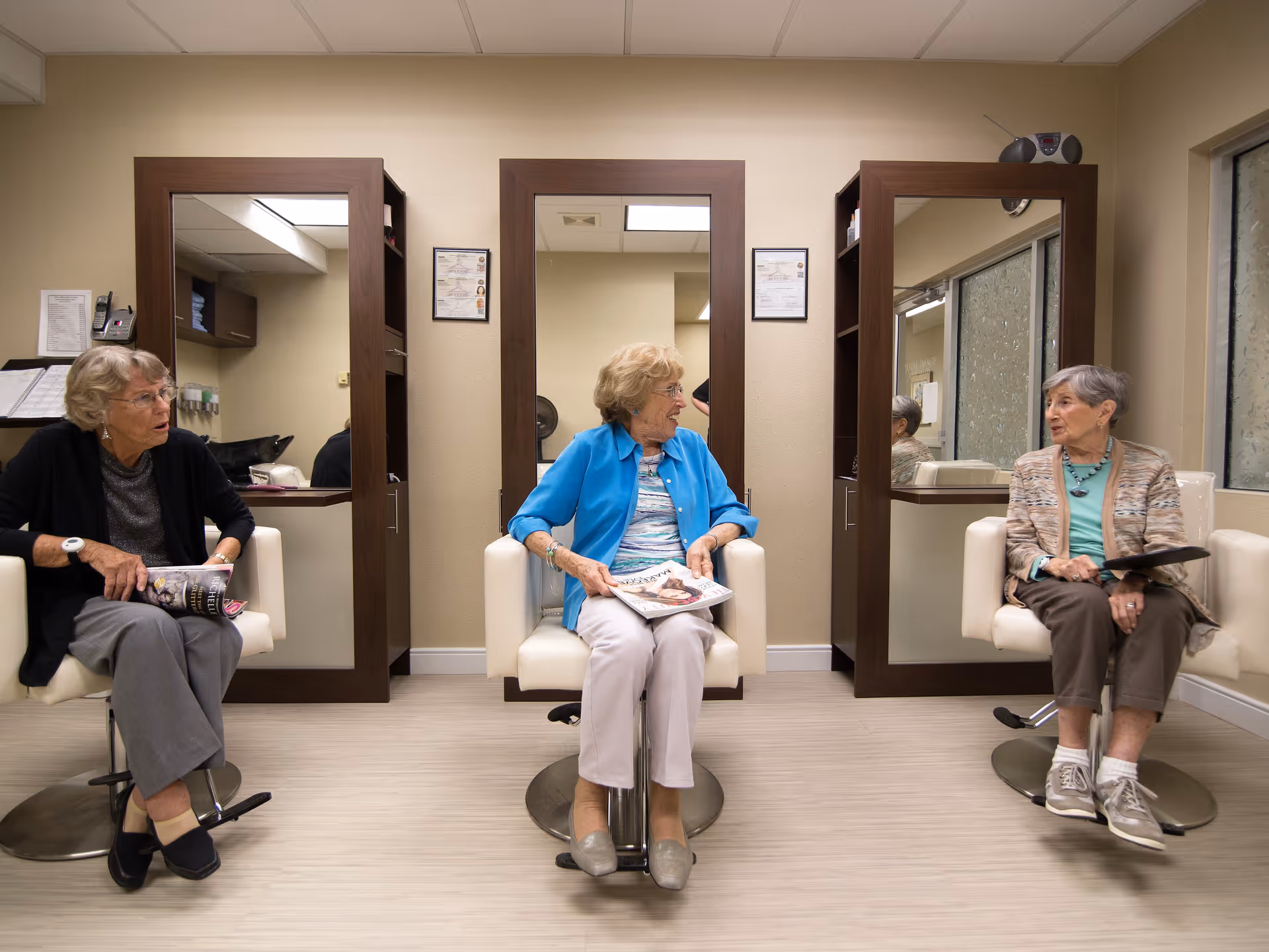 Three elderly women sitting on salon chairs in front of mirrors, holding magazines and engaging in conversation inside a well-lit room with beige walls and wooden framed mirrors.