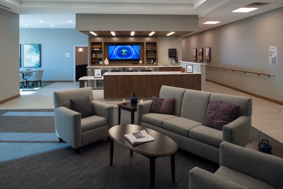 A modern lounge area with a gray sofa and two matching armchairs arranged around a dark wooden coffee table with a newspaper on it. Behind the seating area is a wooden counter with shelves, a mounted flat-screen TV, and a water dispenser. The room has light-colored walls, a carpeted floor in the seating area, and a hallway with handrails on the right side.